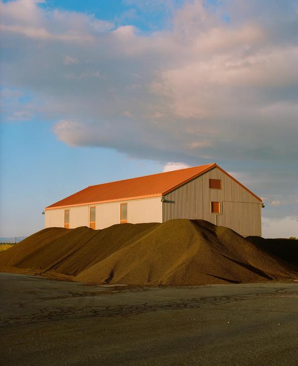 A large industrial building with a red roof stands behind several mounds of dark material under a cloudy sky.