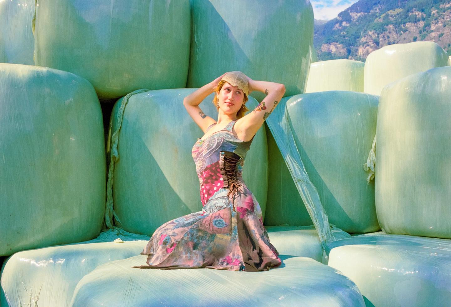 A woman in a patterned dress kneels on a bale of hay wrapped in light green plastic, with mountains in the background.
