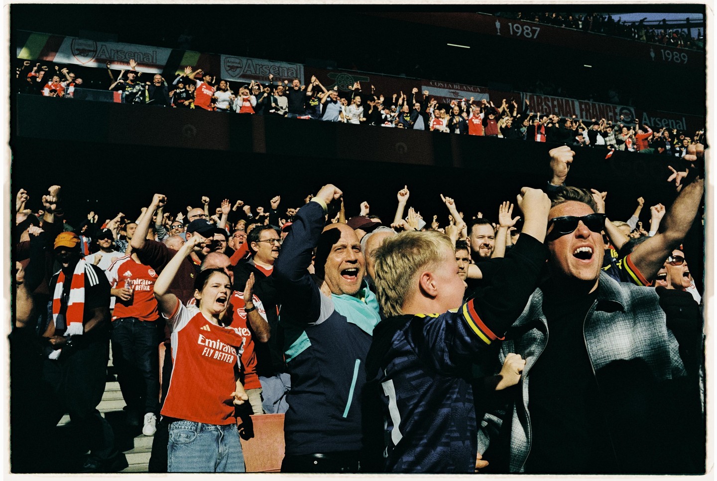 A crowd of excited football fans cheers with their arms raised in a stadium.