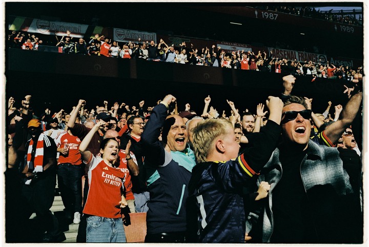 A crowd of excited football fans cheers with their arms raised in a stadium.