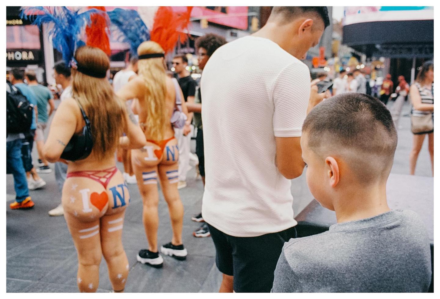 Two women with "I love NY" painted on their buttocks walk through a crowded street.