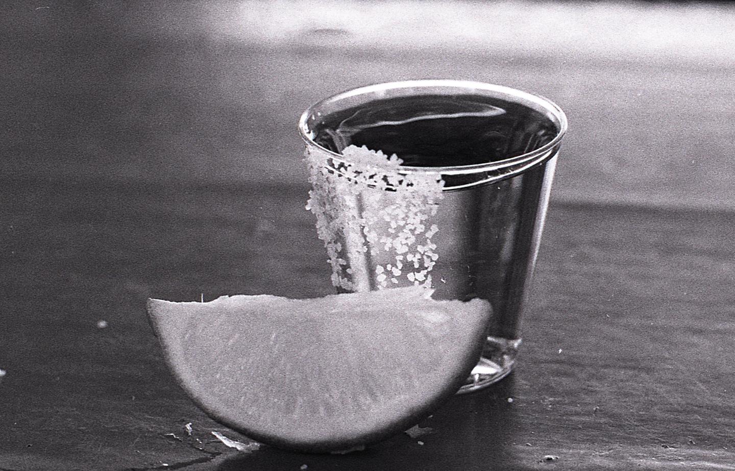 A shot glass with salt on the rim sits next to a lime wedge on a dark surface.