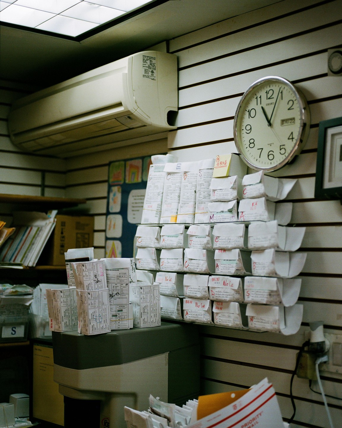 A wall-mounted clock and an air conditioner are visible above shelves filled with small packages in what appears to be a mailroom or office.
