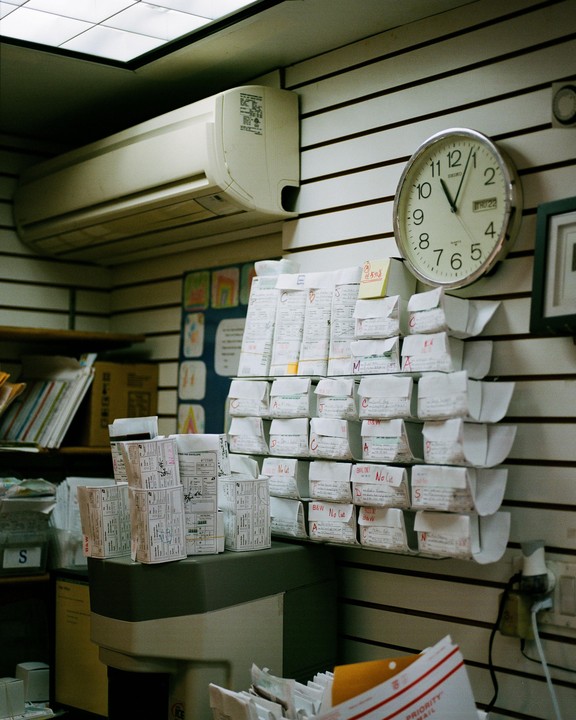 A wall-mounted clock and an air conditioner are visible above shelves filled with small packages in what appears to be a mailroom or office.