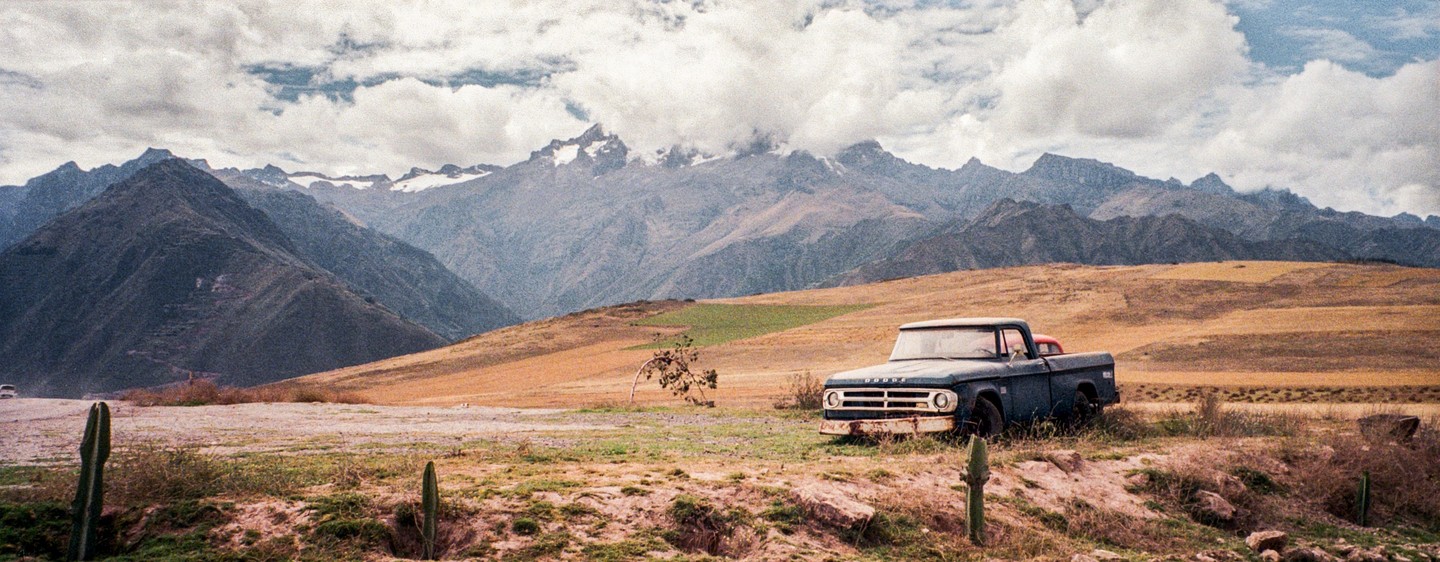 An old blue pickup truck is parked in a field with mountains in the background under a cloudy sky.