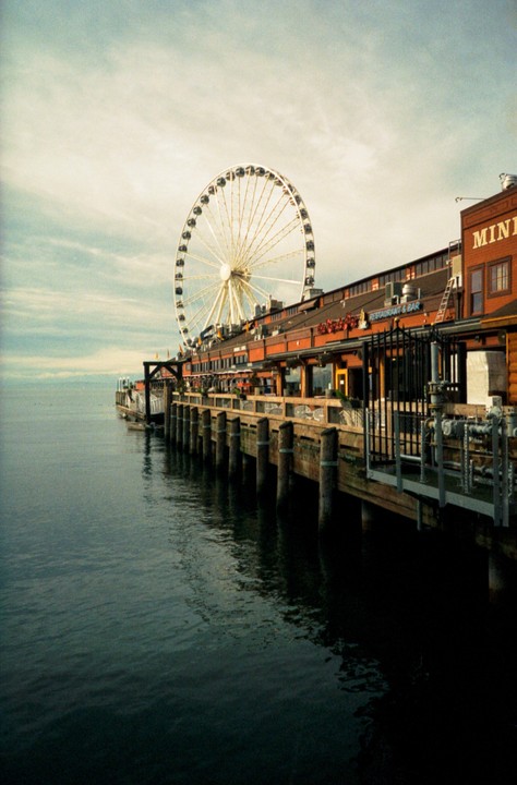 A large Ferris wheel rises above a pier with buildings and water in the foreground.