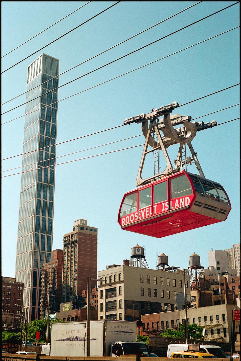 A red Roosevelt Island tram car is suspended in the air above city buildings and power lines.