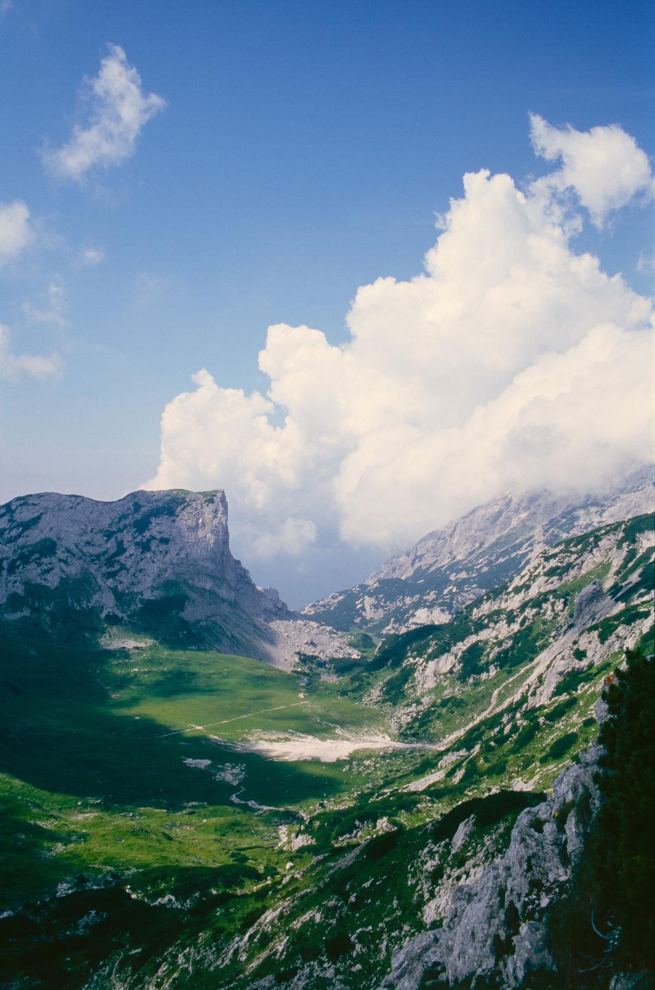 A valley with green grass and rocky mountains is visible under a blue sky with white clouds.