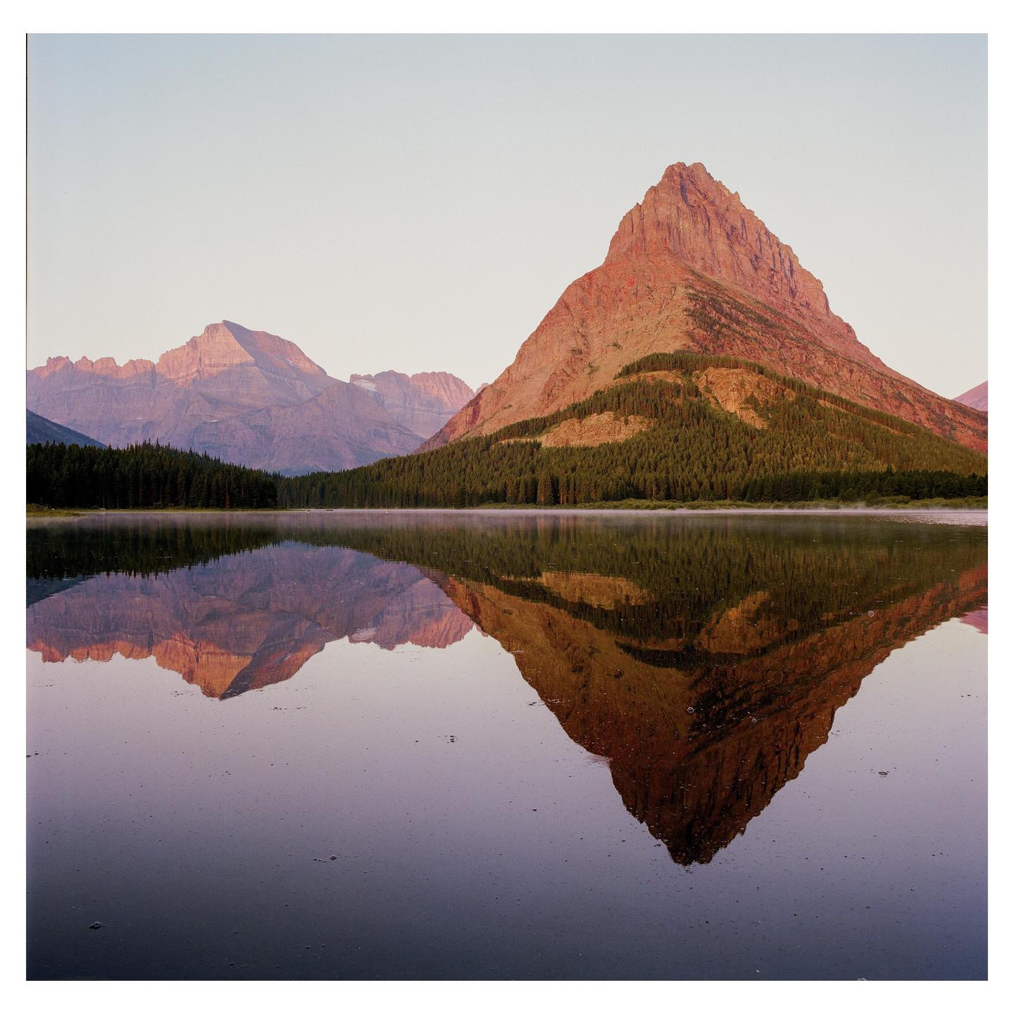 A large mountain with trees on its lower slopes is reflected in a still lake under a clear sky.