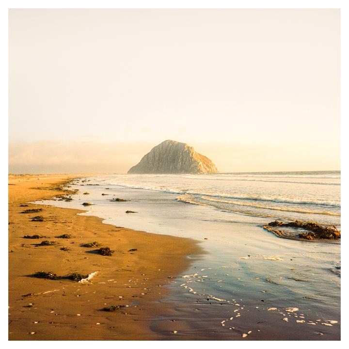 A large rock formation sits in the ocean next to a sandy beach with waves rolling in.