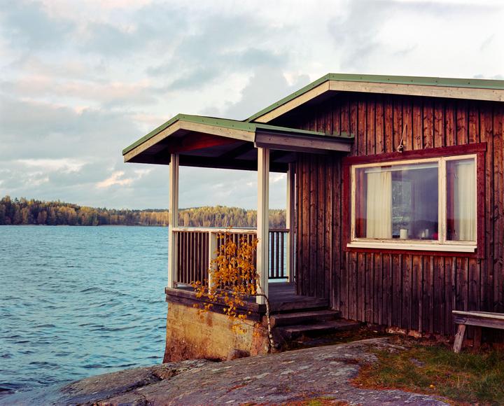 A wooden cabin with a porch sits on a rocky shore next to a lake with a forest in the distance under a cloudy sky.
