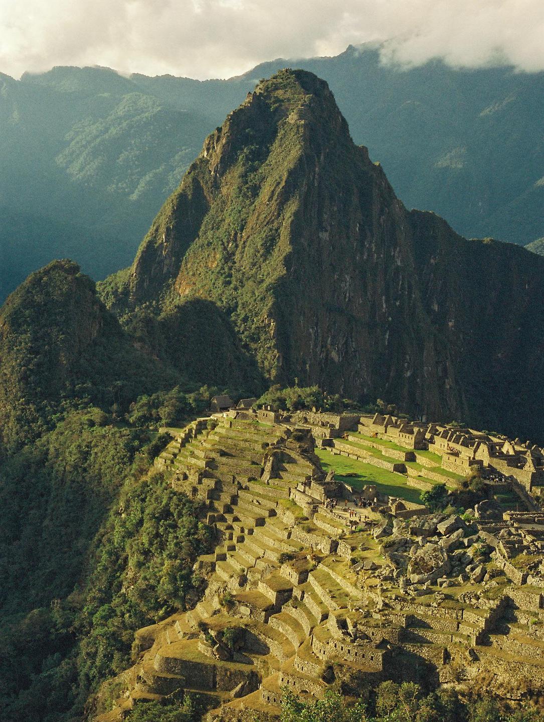 An ancient city with terraced fields is nestled in a valley surrounded by mountains under a cloudy sky.