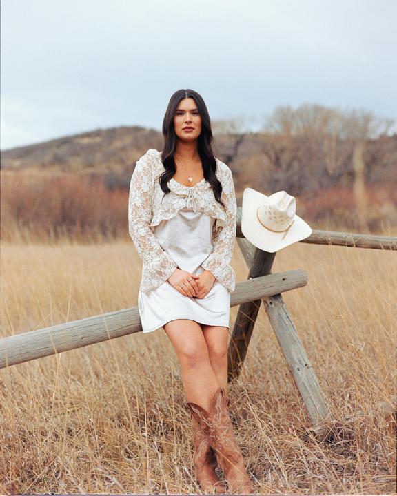 A woman in a white dress and cowboy boots sits on a wooden fence in a field.