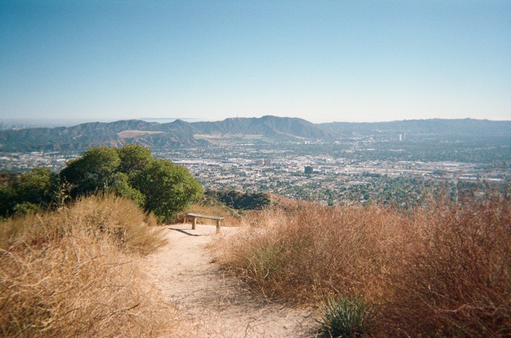A dirt path leads to a wooden bench overlooking a city and mountains under a clear sky.