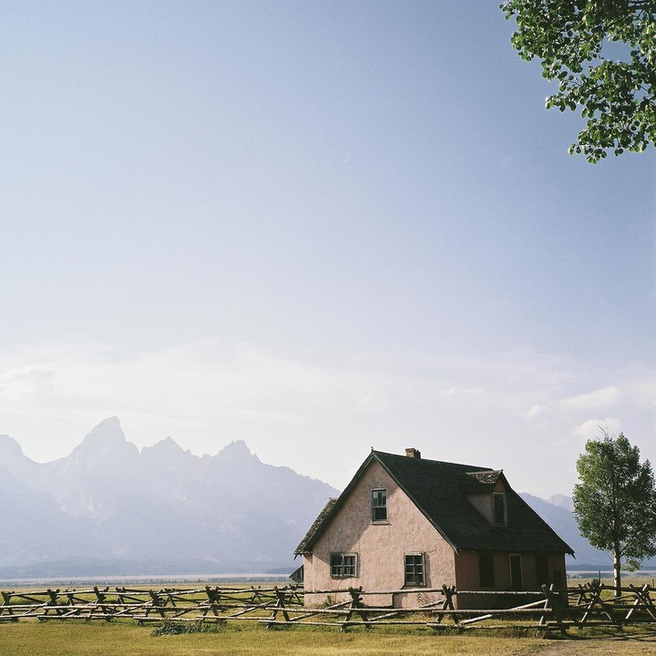 A pink house with a dark roof stands in a field with a wooden fence, with mountains in the background and a tree branch in the foreground.