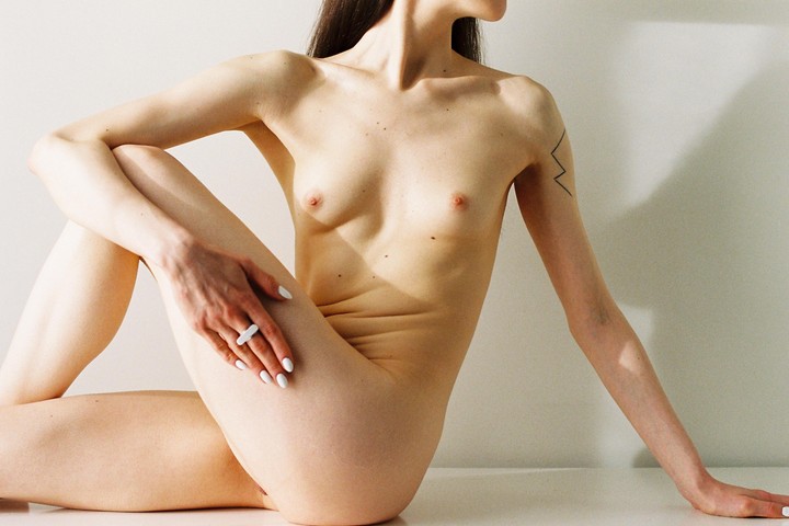 A nude woman with a tattoo on her arm is sitting with her legs crossed and one hand resting on her thigh.