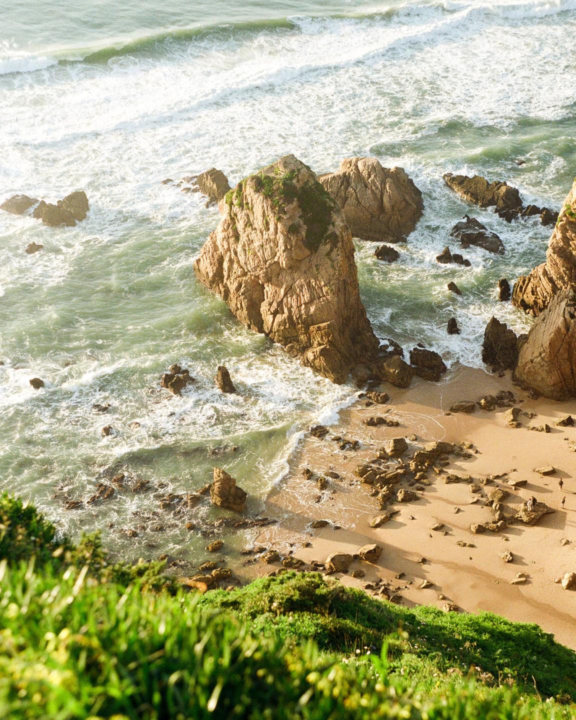 A high-angle view shows a sandy beach with large rocks and ocean waves.