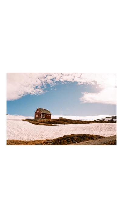 A red house stands on a hill with patches of snow and dry grass under a partly cloudy sky.