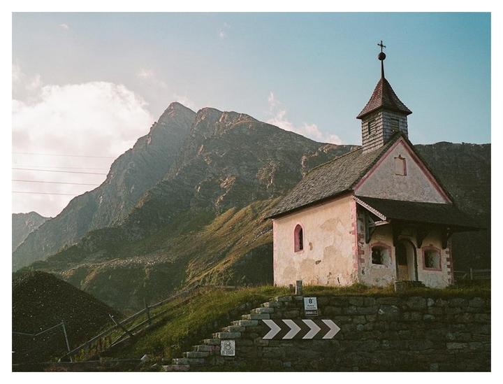 A small chapel with a steeple stands on a hill overlooking a mountain range under a partly cloudy sky.