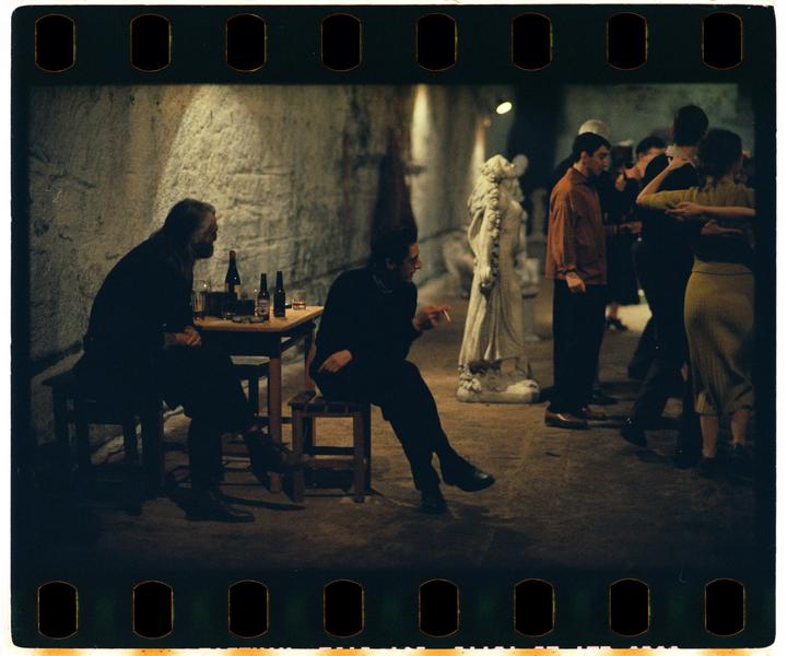 Two men sit at a table with bottles while other people dance in a dimly lit room with a statue.