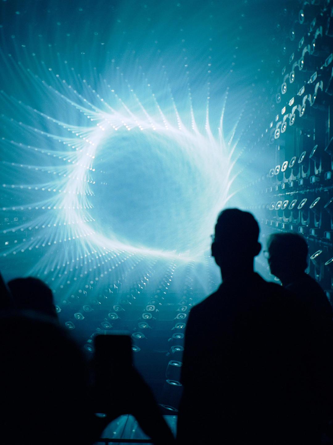 Silhouetted people stand in front of a large blue light installation with a swirling pattern.