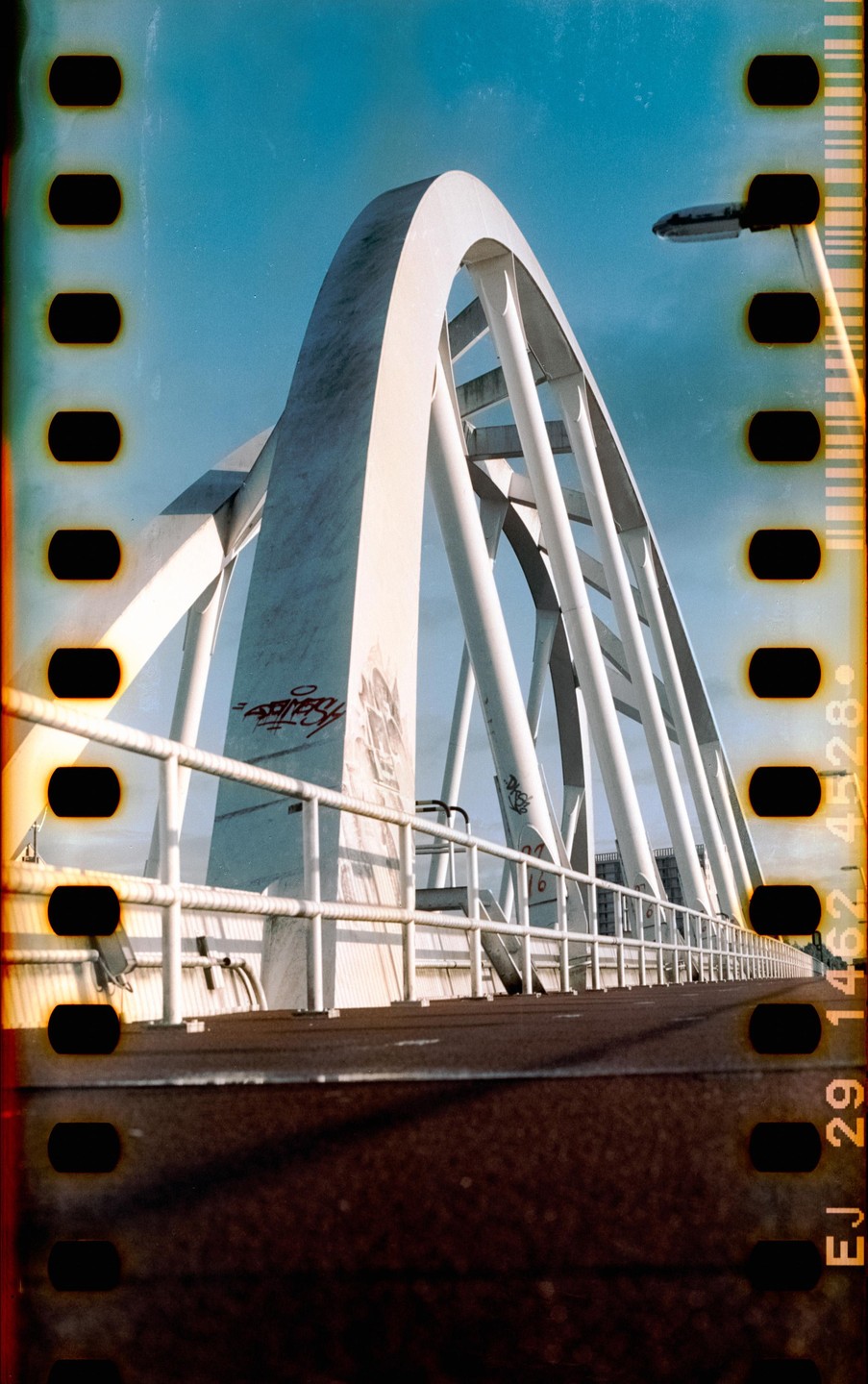 A white arched bridge with graffiti on its supports is viewed from a low angle, with a clear blue sky above.