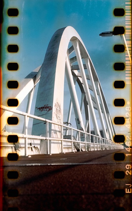 A white arched bridge with graffiti on its supports is viewed from a low angle, with a clear blue sky above.