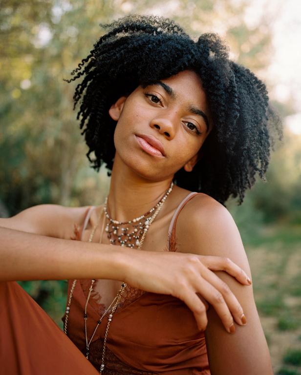 A woman with curly hair and a rust-colored dress looks directly at the viewer.