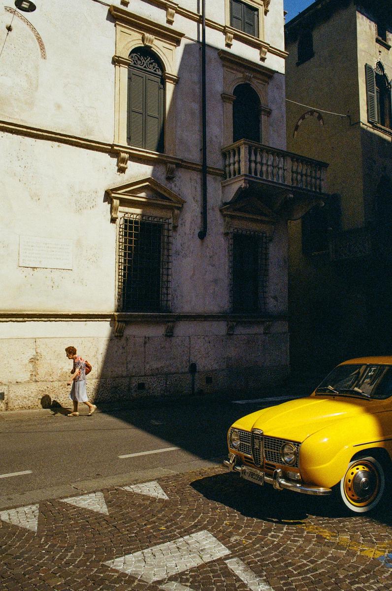 A person walks on a sunlit street next to a yellow car and a building with a balcony.