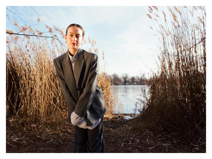 A woman in a suit stands by a lake surrounded by tall, dry reeds.