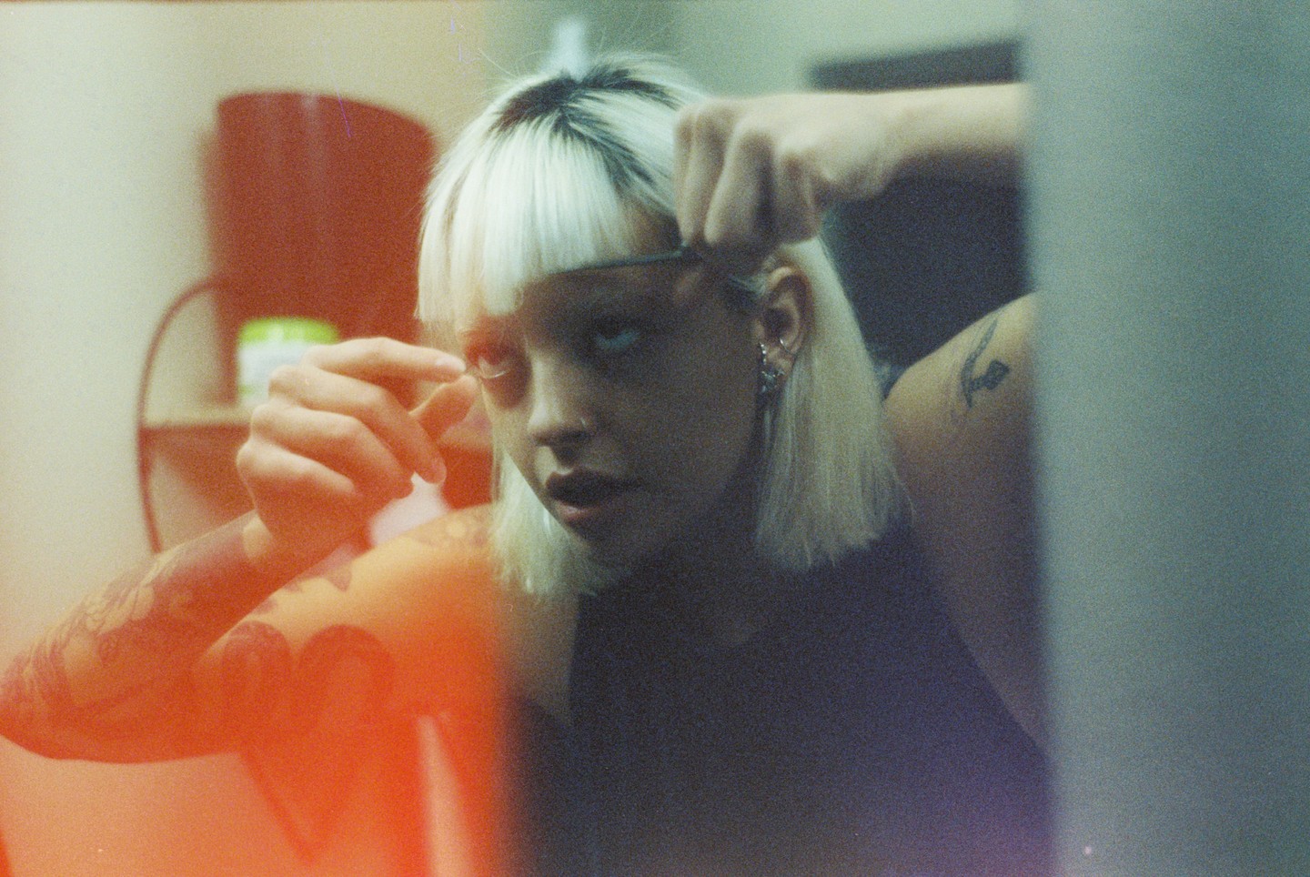 A woman with blonde hair and dark roots applies makeup to her eyelashes while looking into a mirror.