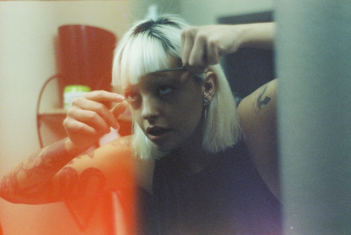 A woman with blonde hair and dark roots applies makeup to her eyelashes while looking into a mirror.
