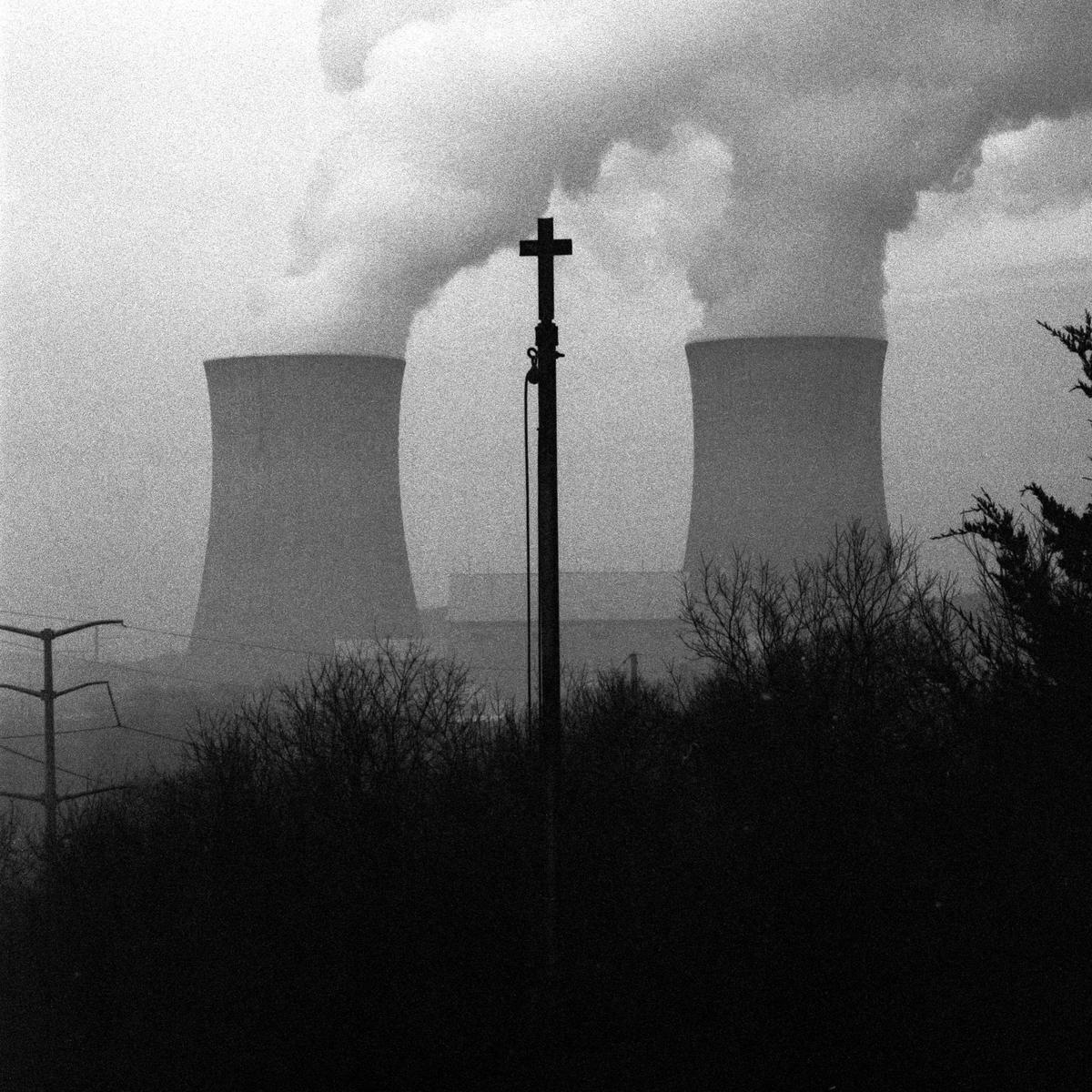 Two cooling towers emit steam behind a cross on a pole and dark bushes.