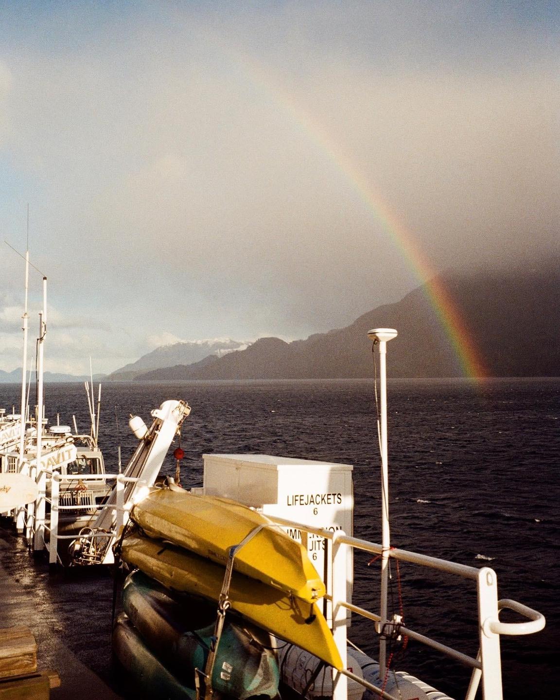 A rainbow stretches over mountains and water as seen from a boat with kayaks on its deck.