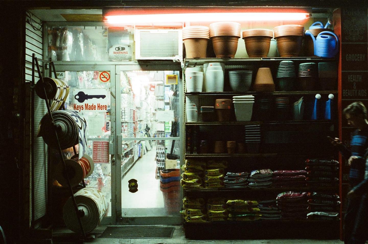 A woman stands outside a brightly lit hardware store at night in New York City.