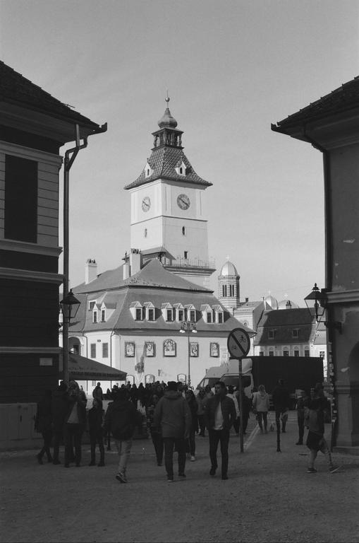 A crowd of people walks on a street toward a large building with a clock tower in Brasov, Romania.