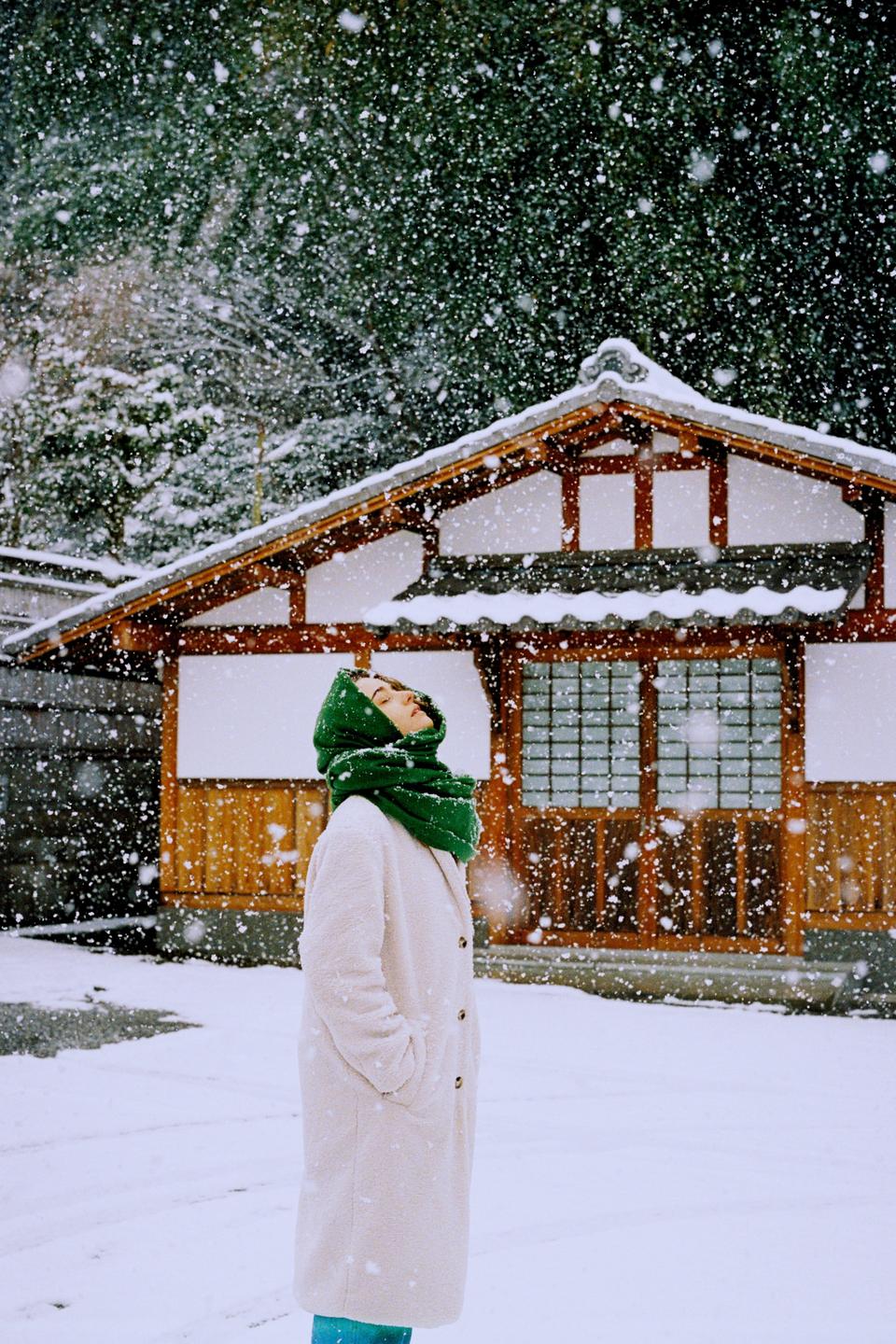 A woman wearing a green scarf and a white coat stands in falling snow, looking up, with a traditional Japanese building in the background.