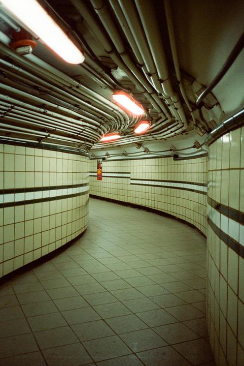 A curved underground tunnel with tiled walls and a ceiling covered in pipes and fluorescent lights is visible.