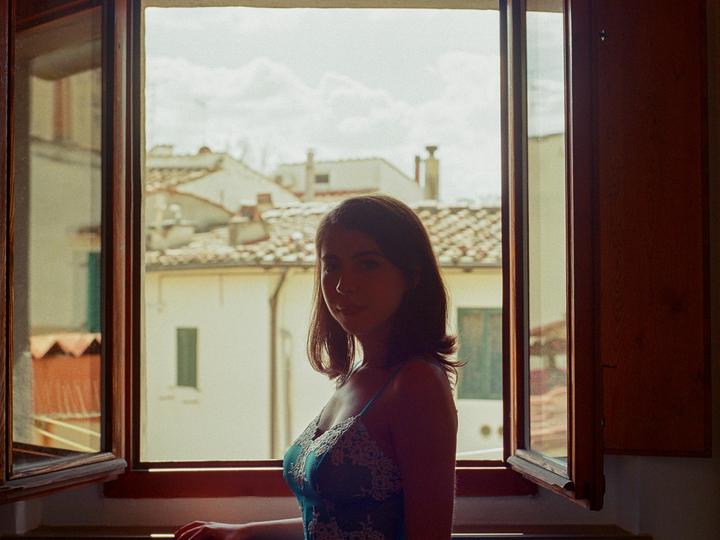 A woman stands in front of an open window looking out at a cityscape with tiled roofs.