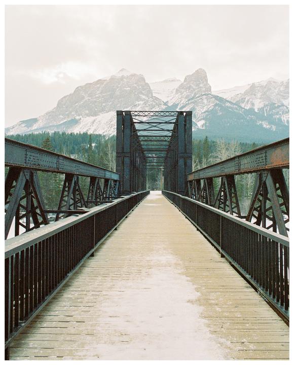 A wooden and metal bridge leads towards snow-capped mountains and a forest under a cloudy sky.