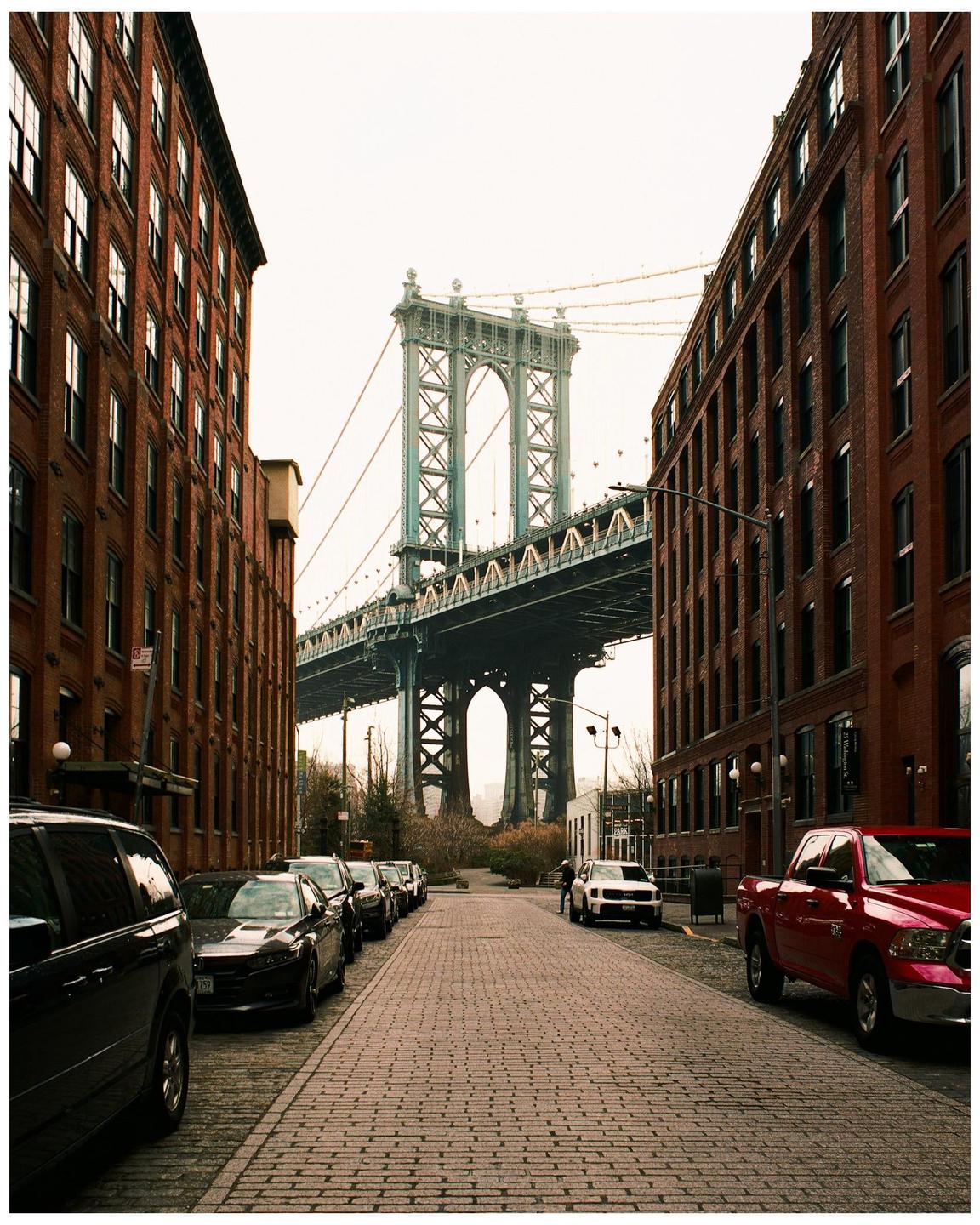 A cobblestone street is lined with parked cars and brick buildings, leading towards a large suspension bridge.