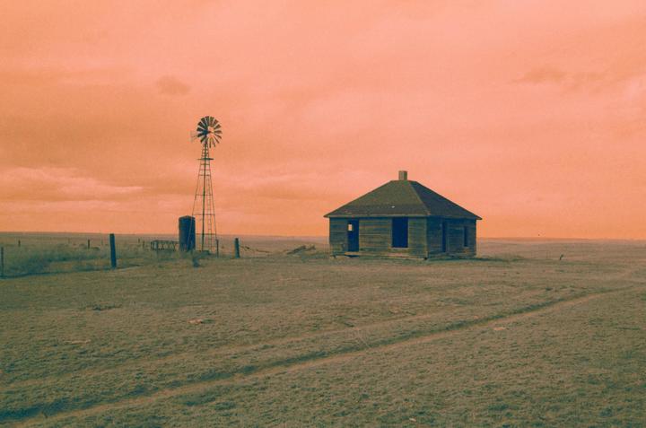 An abandoned house and a windmill stand in a vast, grassy field under an orange sky.