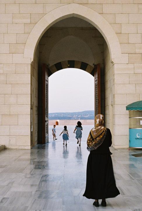 A woman in a headscarf stands in an arched doorway, watching three children run into an open courtyard.