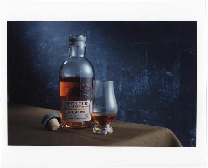 A bottle of Aberlour whisky, a glass with whisky, and a cork are arranged on a brown tablecloth against a dark blue background.