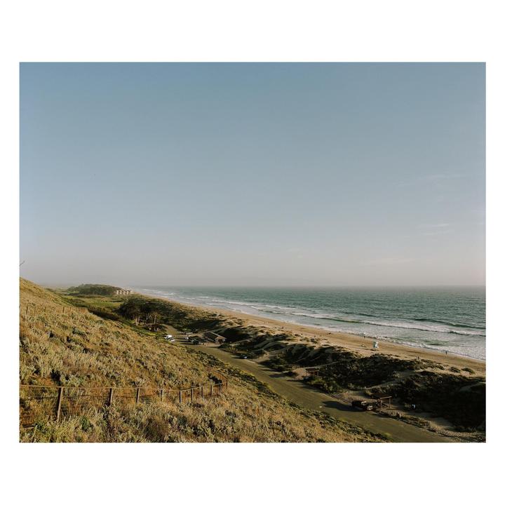 A long stretch of beach and ocean is visible from a grassy hillside.