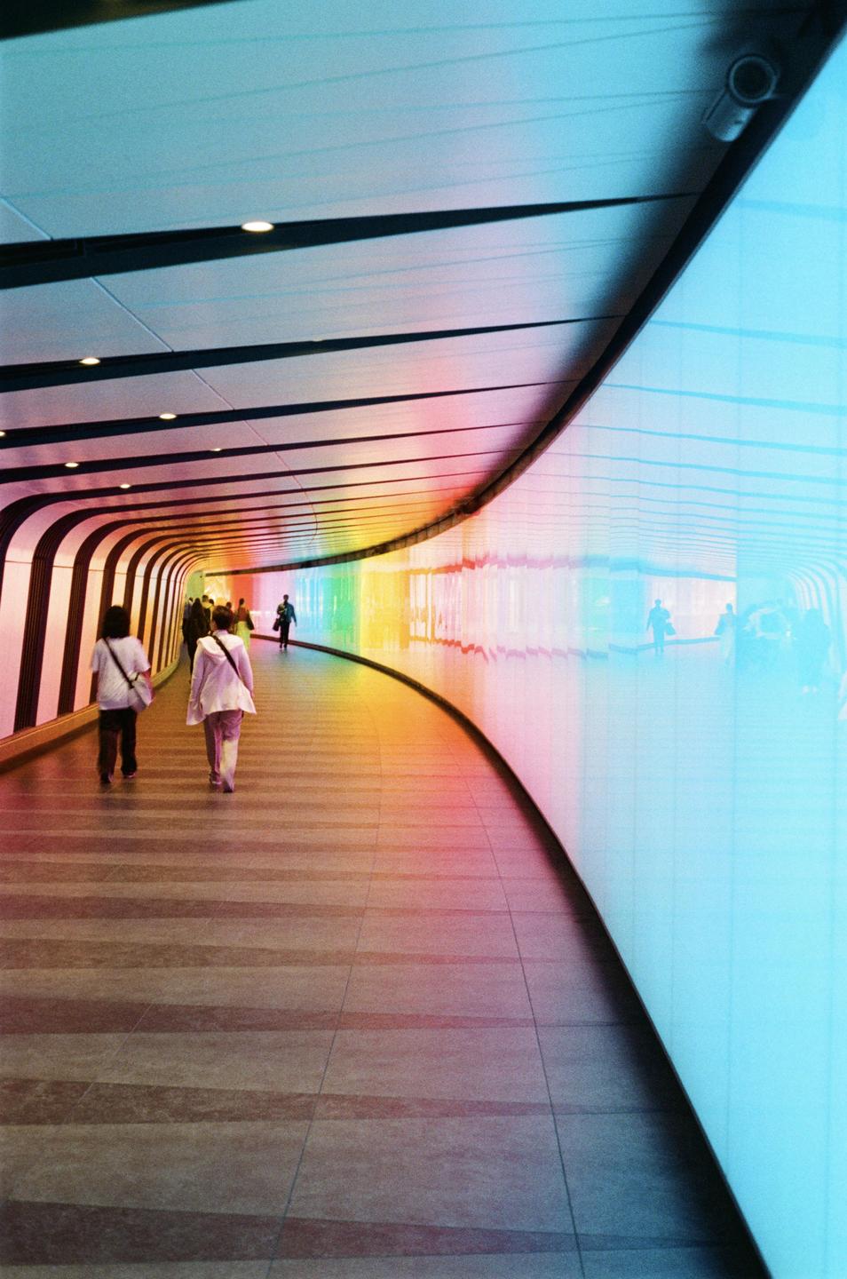 People walk through a curved tunnel with rainbow-colored lights on the wall.