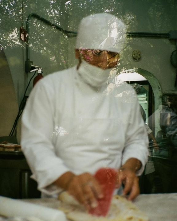 A chef in a white uniform is preparing food with a red tool, seen through a window with a reflection of trees.