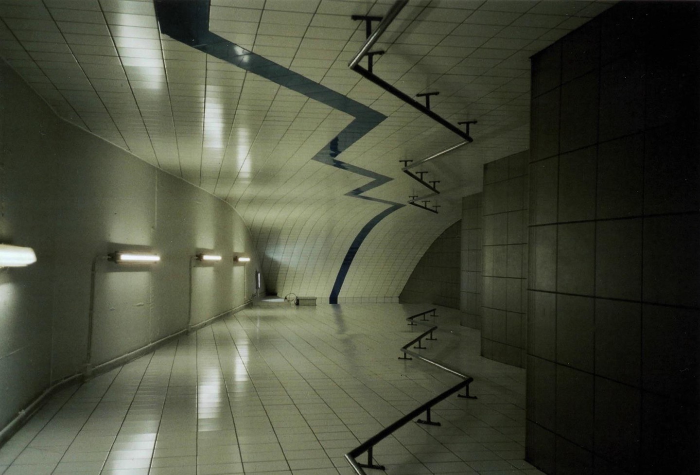 A long, tiled hallway with a zigzagging blue line on the ceiling and a series of metal rails on the floor.