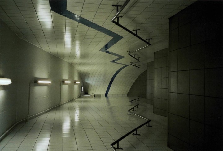 A long, tiled hallway with a zigzagging blue line on the ceiling and a series of metal rails on the floor.