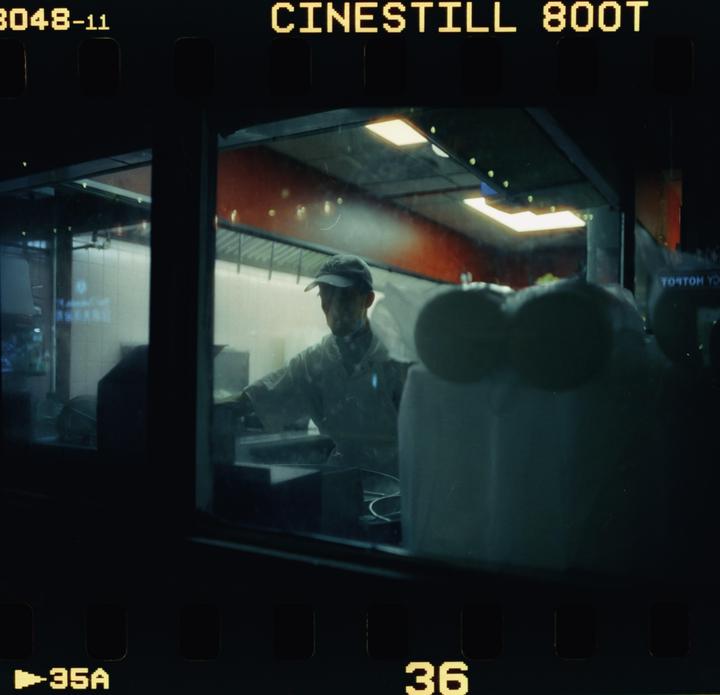 A man in a cap and white shirt works in a brightly lit kitchen at night, viewed through a window.